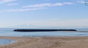 A beach with a rocky breakwater extending into calm waters, a sandy shore, and distant mountains under a clear sky.