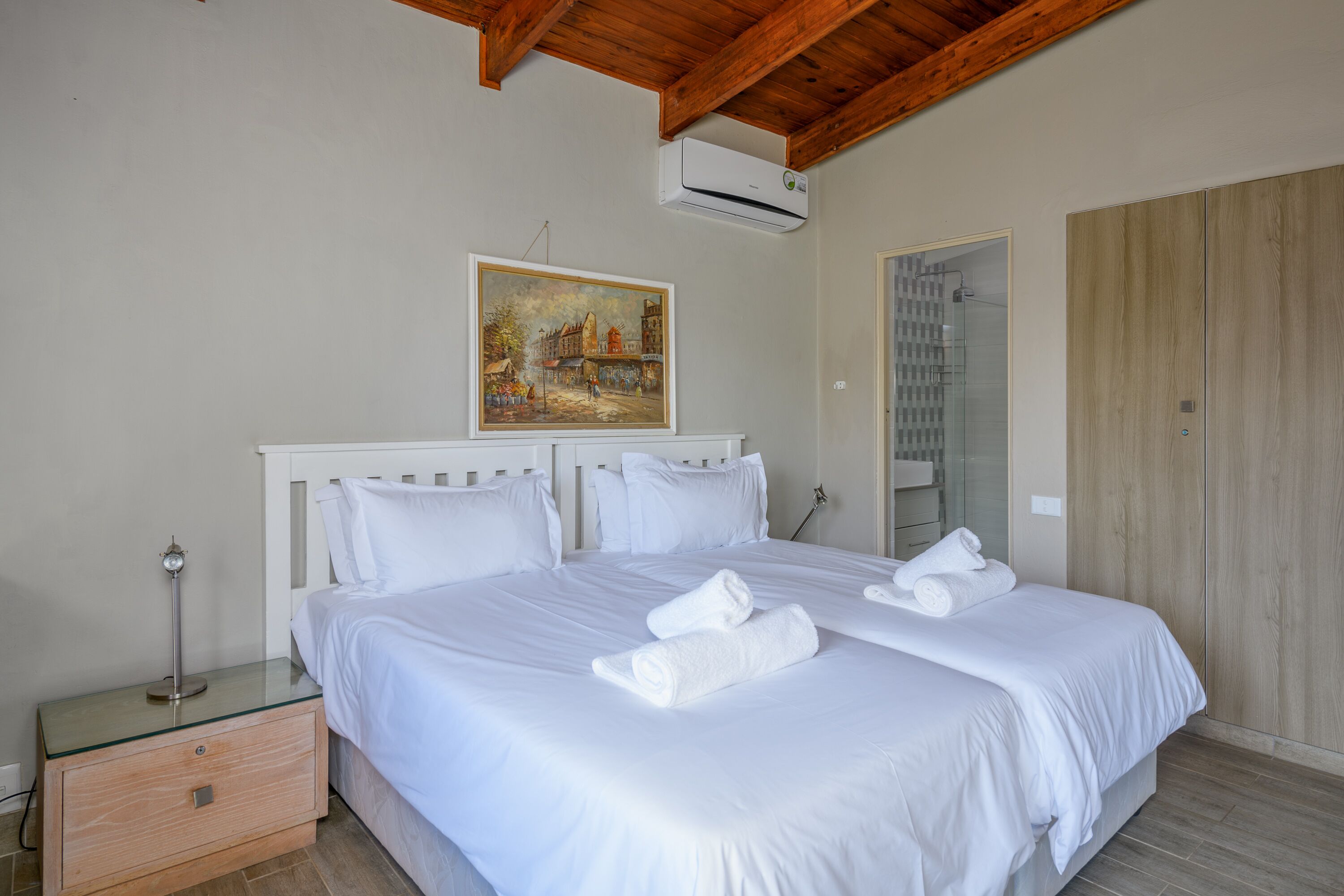 Una cama bien hecha con ropa de cama y toallas blancas, una mesita de noche con lámpara, un cuadro en la pared y un armario de madera.