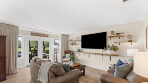 A living room with a sofa, armchair, coffee table, and a flat-screen TV mounted on the wall. There are shelves with decorative items and a sign that reads "BEACH HOUSE".