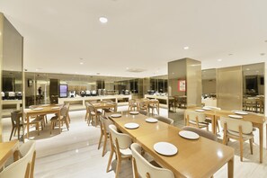 A modern dining area with multiple tables and chairs, a kitchen in the background, and a clean, well-lit environment. - Ji Hotel (Shanghai)