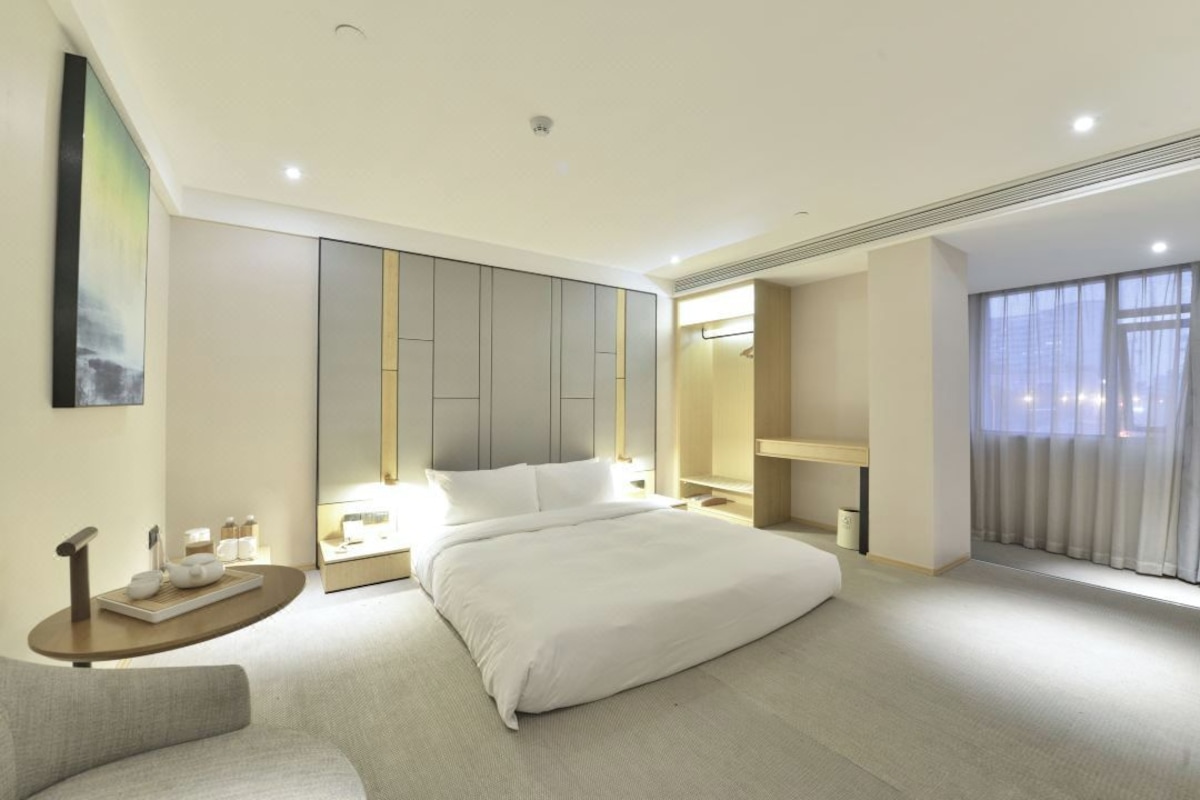 A modern hotel room with a large bed, bedside tables, a seating area, and a large window with curtains.