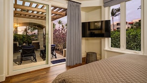 Ein Schlafzimmer mit einem großen Bett, einem an der Wand befestigten Fernseher und einer Glastür, die auf eine Terrasse im Freien mit Möbeln und einem Kinderwagen führt.