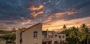 Um edifício moderno com telhado plano, várias varandas e um jardim bem cuidado, com palmeiras e arbustos.