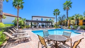 A poolside area with lounge chairs and a table, surrounded by palm trees and a building with a tiled roof. - Golf Carts ~ Patio ~ Balcony ~ Community Pool (Miramar Beach)