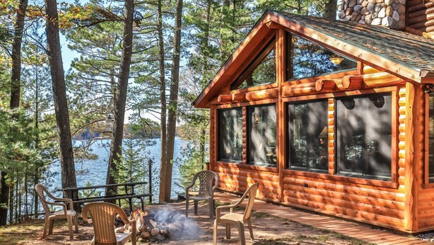 Une cabane en rondins dotée de grandes fenêtres donnant sur un lac, entourée d’arbres.