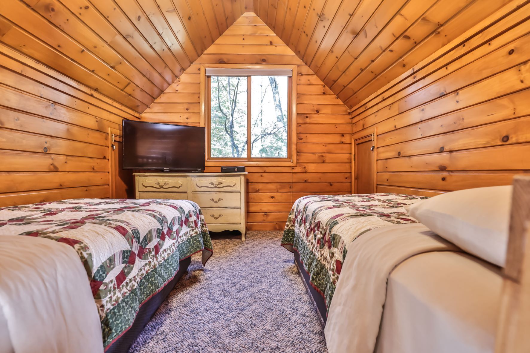 A cozy cabin room with two beds, a dresser, a television, and a window with a view of trees.