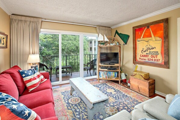 A living room with a red sofa, a coffee table, a TV, and a bookshelf. There is a large window with a view of trees and a balcony. - Oceanfront Condominium with Great Amenities (Saint Simons Island)