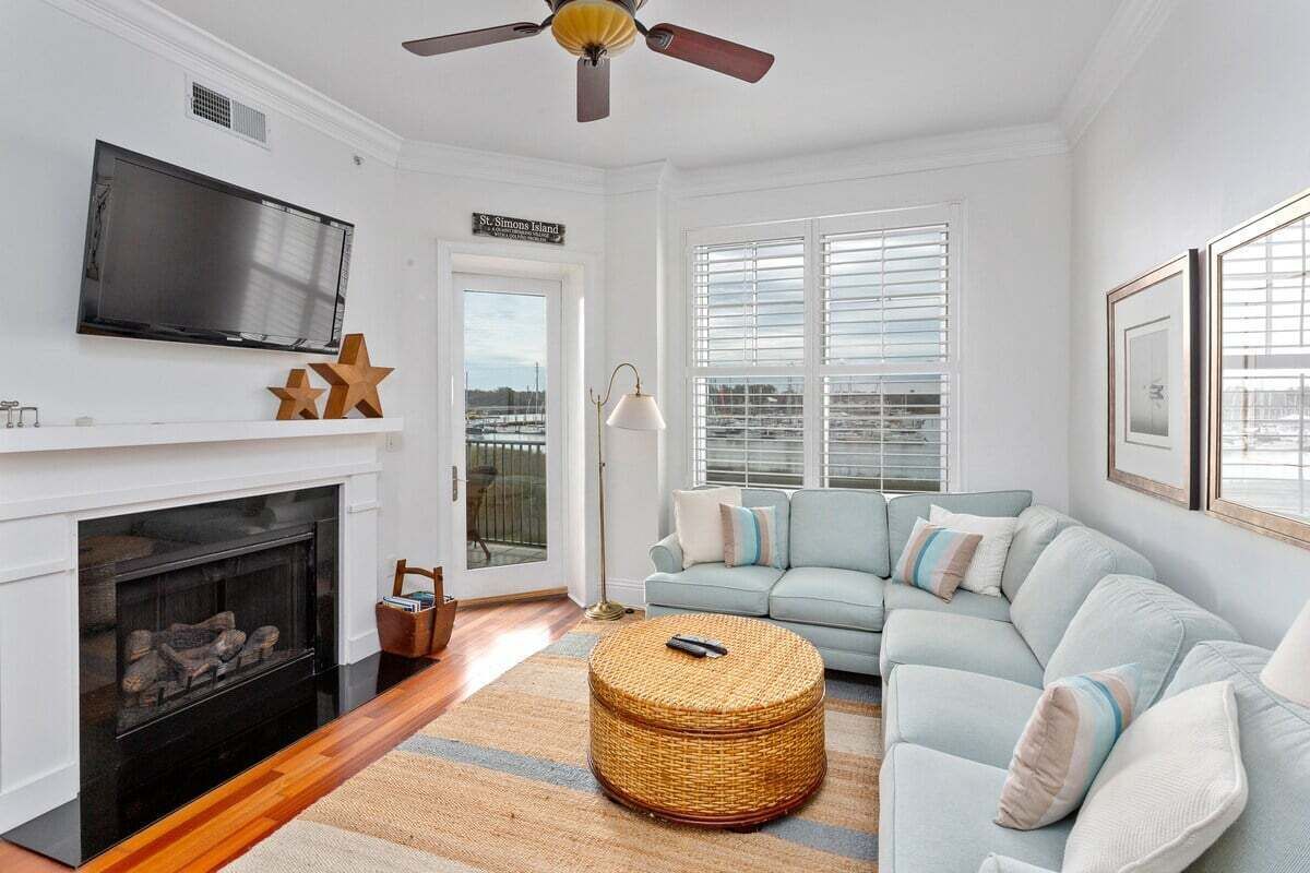 A living room with a fireplace, a television, a light-colored sofa, and a large window with blinds.