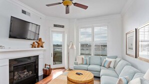 A living room with a fireplace, a television, a light-colored sofa, and a large window with blinds. - Breathtaking Views of the Inter-coastal Waterway (Saint Simons Island)