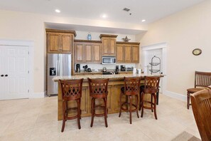A kitchen with wooden cabinets, a stainless steel refrigerator, a microwave, and a coffee maker. There are four wooden bar stools at the island. - Sea Point Condo #306 (Saint Simons Island)