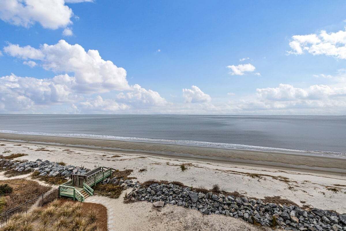 A beach with a wooden staircase leading to a small structure, a rocky barrier, and a clear view of the ocean.