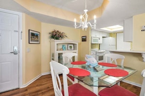 Uma área de refeições com mesa de vidro, jogos americanos vermelhos, cadeiras brancas, um candelabro e uma cozinha ao fundo.