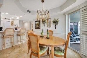 A dining area with a round wooden table, four chairs with green cushions, a chandelier, and a view of the outside through a sliding glass door. - Family Friendly Direct Ocean View Condo (Saint Simons Island)