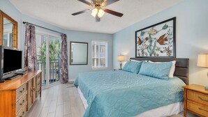 A bedroom with a large bed, a ceiling fan, a television, a dresser, and a balcony with curtains. - Oceanfront Condominium with Great Amenities (Saint Simons Island)