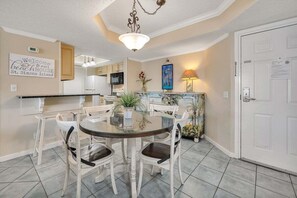 A dining area with a round table and white chairs, a kitchen in the background, and a sign on the wall welcoming visitors to the beach house. - St Simons Oceanfront Condominium (Saint Simons Island)