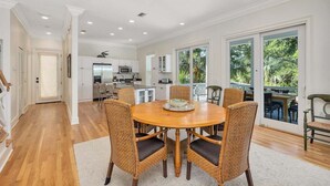 Uma sala de jantar ampla com uma mesa redonda de madeira e cadeiras de vime, uma cozinha ao fundo, e grandes portas de vidro deslizantes que dão para uma varanda com vista para o verde.