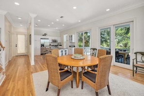 A spacious dining area with a round wooden table and wicker chairs, a kitchen in the background, and large sliding glass doors leading to a balcony with a view of greenery.