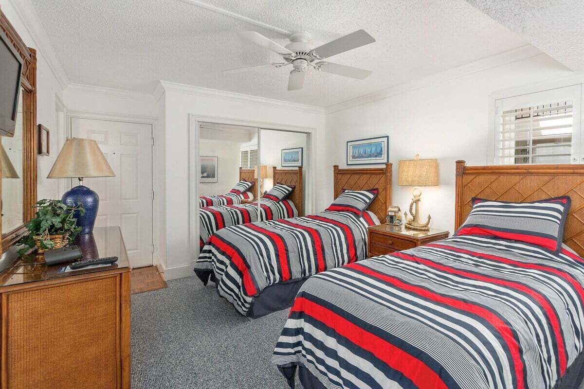 A hotel room with three beds, a ceiling fan, a desk with a lamp, and a window with blinds.