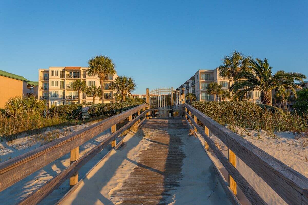 A wooden boardwalk leading to a beachfront property with multiple-story buildings, palm trees, and a clear sky.