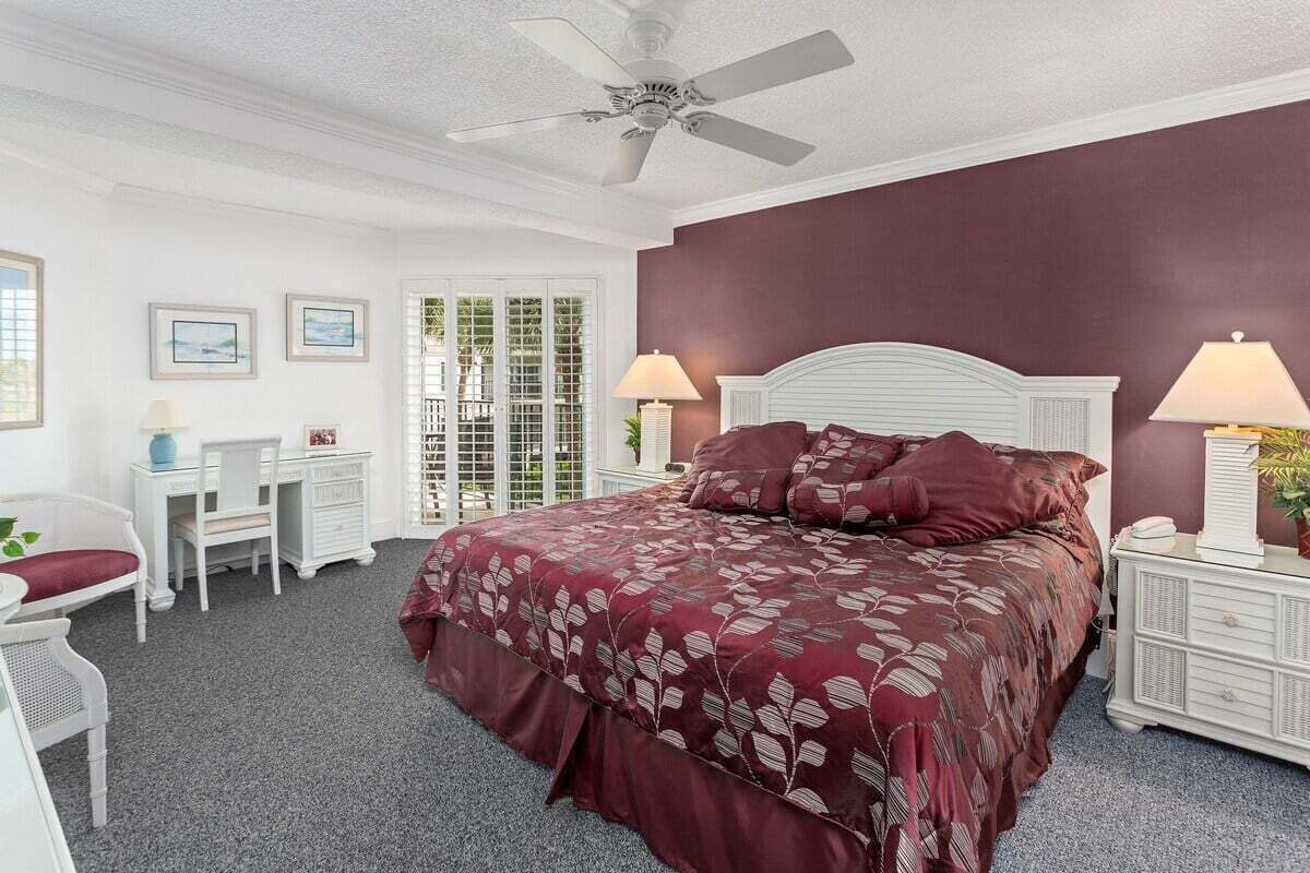 A bedroom with a large bed, two bedside tables with lamps, a white ceiling fan, and a window with blinds.