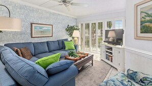 A living room with a blue sofa, a ceiling fan, a TV on a white cabinet, and a window with blinds. - Steps from the Beach! (Saint Simons Island)