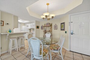 A dining area with a glass table, white wicker chairs, a chandelier, and a kitchen in the background. - Oceanfront Condominium with Great Amenities (Saint Simons Island)
