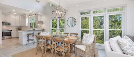 A spacious kitchen with white cabinetry, a dining area with a round table and chairs, and a large window offering a view of greenery.