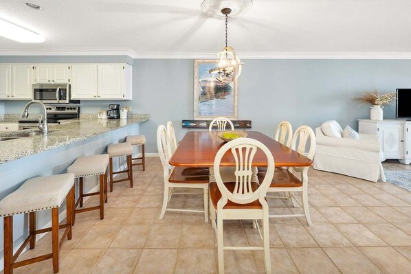 A spacious dining area with a wooden table and white chairs, a kitchen with white cabinets, and a ceiling fan. - Oceanfront Condo in the Heart of The Village. (Saint Simons Island)