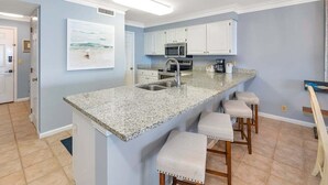 A kitchen with a granite countertop, white cabinets, a stainless steel sink, and a microwave. There are three bar stools with cushioned seats. - Oceanfront Condo in the Heart of The Village. (Saint Simons Island)