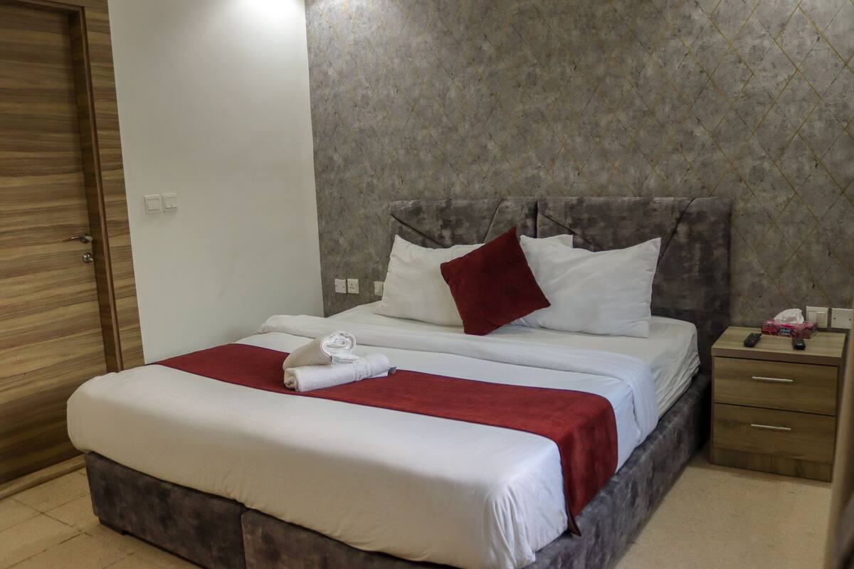 Una cama bien tendida con ropa de cama blanca y roja, una mesita de noche de madera con un control remoto y una pared con estampado de mármol.