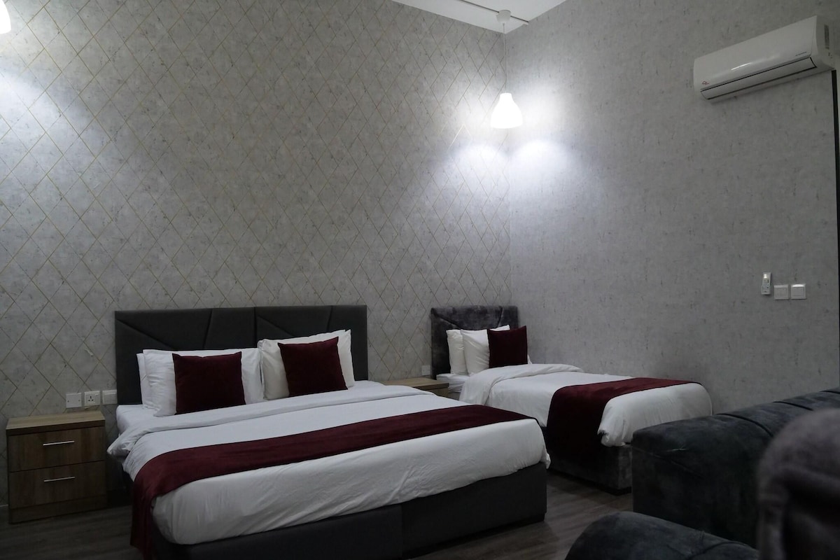 Una habitación de hotel con dos camas, un cabecero oscuro, una mesita de noche de madera, un sofá gris y un equipo de aire acondicionado en la pared.