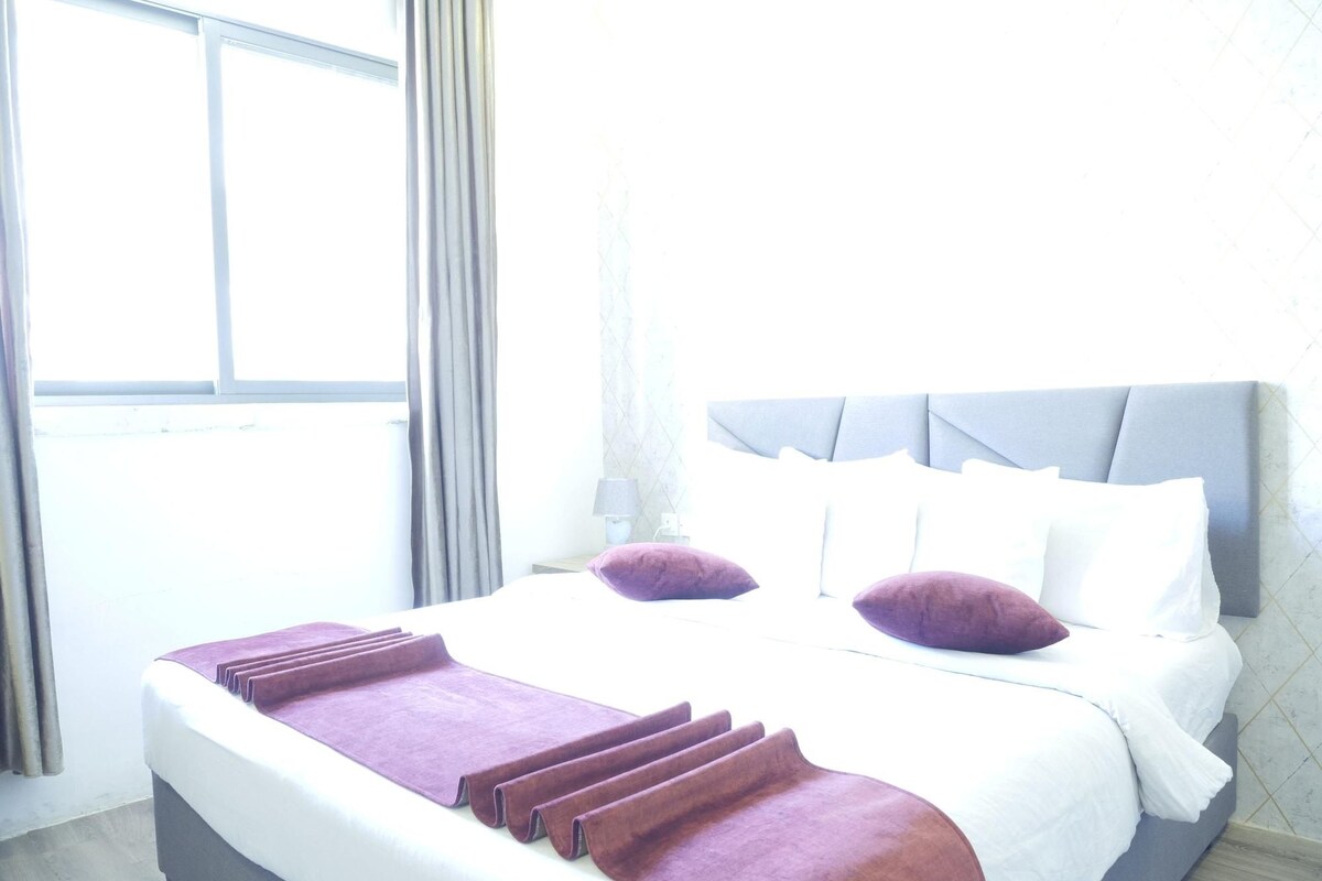 A hotel room with a large bed, grey headboard, and a window with curtains.