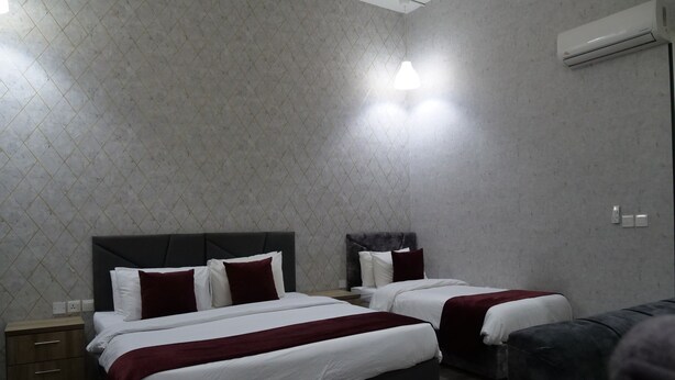 Une chambre d’hôtel avec deux lits, une tête de lit sombre, une couvre-lit rouge et blanche, un canapé gris et un climatiseur fixé au mur.