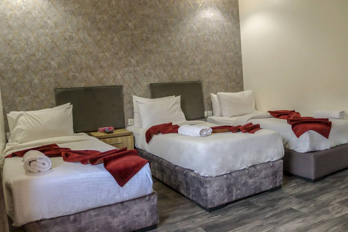 Habitación de hotel con tres camas, cada una con ropa blanca y mantas rojas. La habitación tiene un papel tapiz estampado y piso de madera.