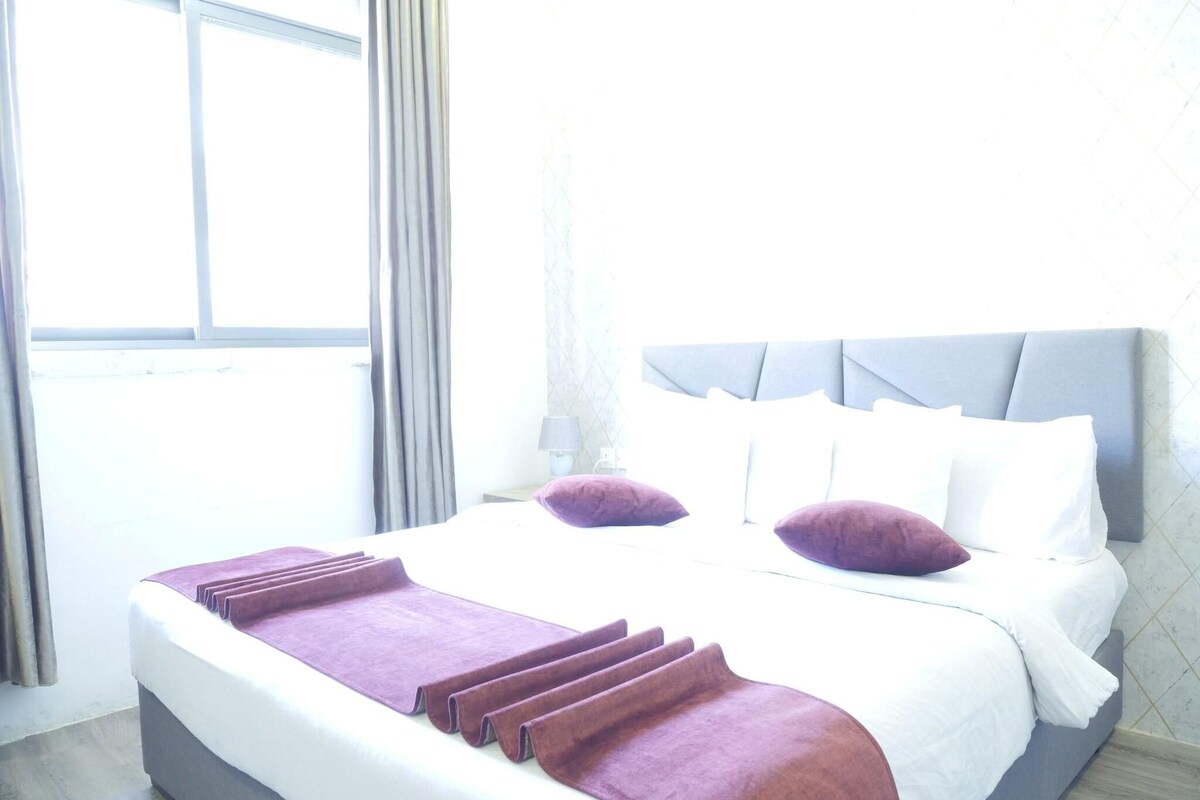 A hotel room with a large bed, grey headboard, and a window with curtains.