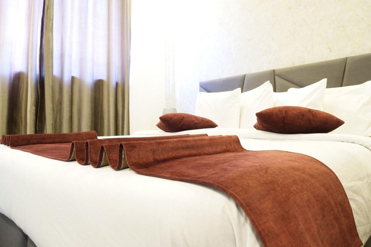 Una cama bien hecha con sábanas blancas y almohadas marrones, un cabecero gris y una ventana con cortinas.