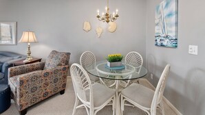 A dining area with a glass table, white wicker chairs, a patterned sofa, a chandelier, and a sailboat painting on the wall.
