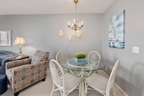 A dining area with a glass table, white wicker chairs, a patterned sofa, a chandelier, and a sailboat painting on the wall.