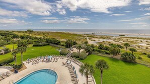 A pool area with lounge chairs and umbrellas, surrounded by greenery and a view of the beach and ocean. - Vacation on St Simons Island! Pool, Beach Access, Fitness Center, Tennis! (Saint Simons Island)