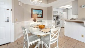 A dining area with a round table, white chairs, a chandelier, and a kitchen in the background. - Vacation on St Simons Island! Pool, Beach Access, Fitness Center, Tennis! (Saint Simons Island)