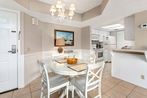 A dining area with a round table, white chairs, a chandelier, and a kitchen in the background.