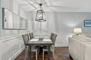 A dining area with a rectangular table, four chairs, a hanging light fixture, a mirror, a wall-mounted lamp, and a framed picture. - Oceanfront Condominium with Balcony and Pool (Saint Simons Island)