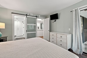 A bedroom with a bed, a dresser, a television, and a bathroom with a shower and toilet. - Oceanfront Condominium with Balcony and Pool (Saint Simons Island)