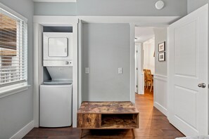 A compact laundry room with a built-in washer and dryer, a small wooden bench, and a door leading to a dining area. - Oceanfront Condominium with Balcony and Pool (Saint Simons Island)