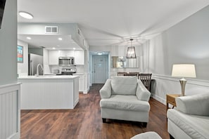 A modern kitchen with white cabinetry, a central island, and a dining area with a white sofa and table. - Oceanfront Condominium with Balcony and Pool (Saint Simons Island)