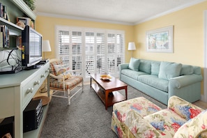 Relax in this inviting living room with a sofa, ottoman, and armchair, perfect for unwinding near a vintage TV and a beautiful window.