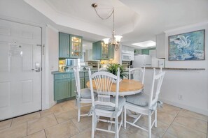 A dining area with a round wooden table and white chairs, a chandelier, and teal cabinetry in the background. - Golden Isles Spacious and Oceanfront Condo (Saint Simons Island)