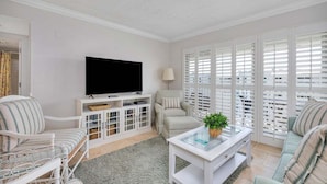 Un salón con televisor de pantalla plana, muebles blancos, una mesa de centro de vidrio y vistas amplias a través de ventanas con contraventanas.