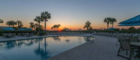 A swimming pool with lounge chairs and umbrellas, palm trees, and a sunset view.