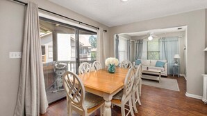 A dining area with a wooden table and white chairs, a glass door leading outside, and a living room with a sofa and curtains. - Enjoy being steps from the Pier Village at #508! (Saint Simons Island)